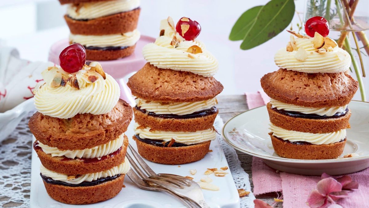 Auf einem Tisch sind 3 Frankfurter-Kranz-Cupcakes angerichtet. Sie bestehen aus drei Schichten mit Buttercreme und Kuvertüre. Obendrauf ist ein Tüpfel Buttercreme und jeweils eine Kirsche.