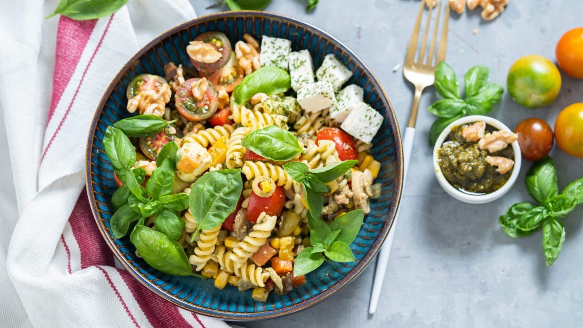 Unser italienischer Pasta Salat angerichtet in einer Schüssel. Im Hintergrund sind Tomaten, Pesto und Walnüsse zu sehen.