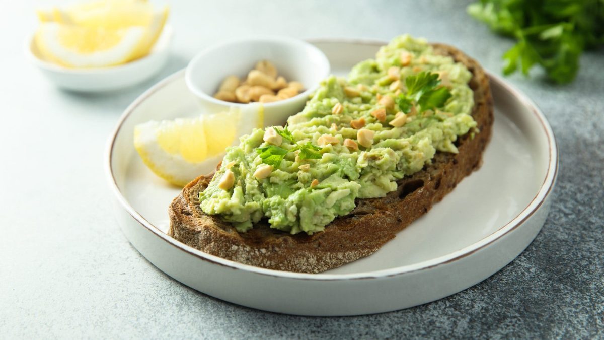 Unser Guacamole-Brot liegt angerichtet mit Erdnüssen und Zitronenschnitzen auf einem weißen Teller.