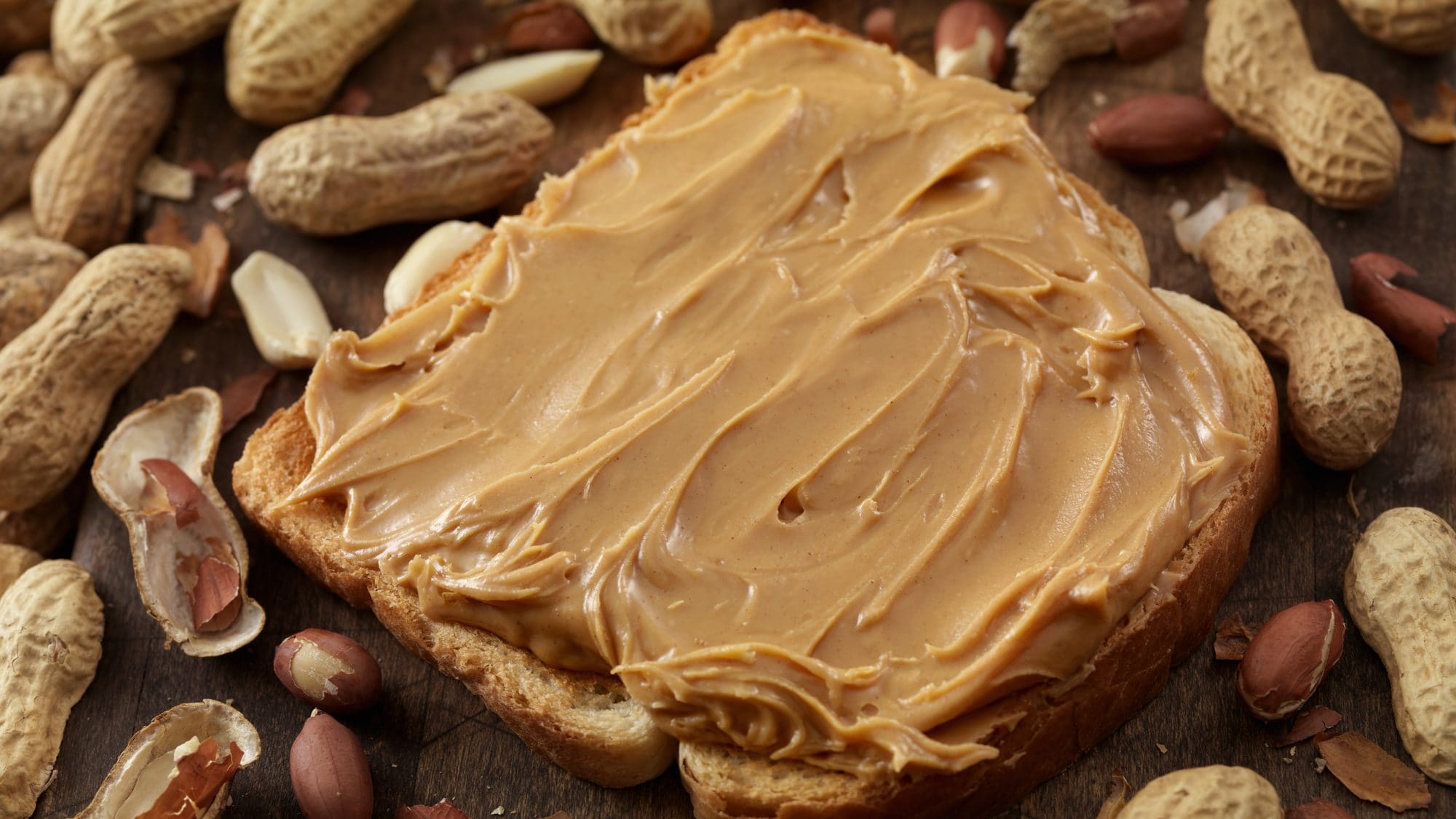 Eine Scheibe Erdnussbutter-Toast liegt inmitten von zahlreichen Erdnüssen auf dem Tisch.