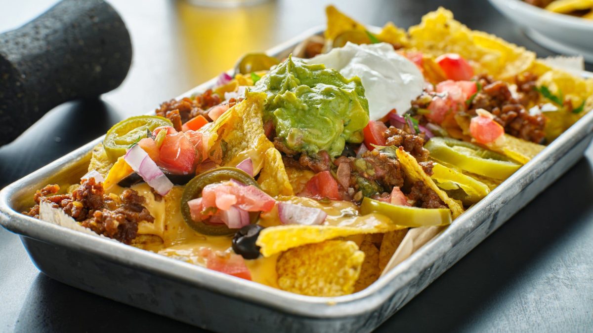 Loaded Nachos mit Chili in weißer Auflaufform auf grauem Untergrund. Links angedeutet dunkelgrauer Mörser und Olivenöl in Karaffe. Frontalansicht.