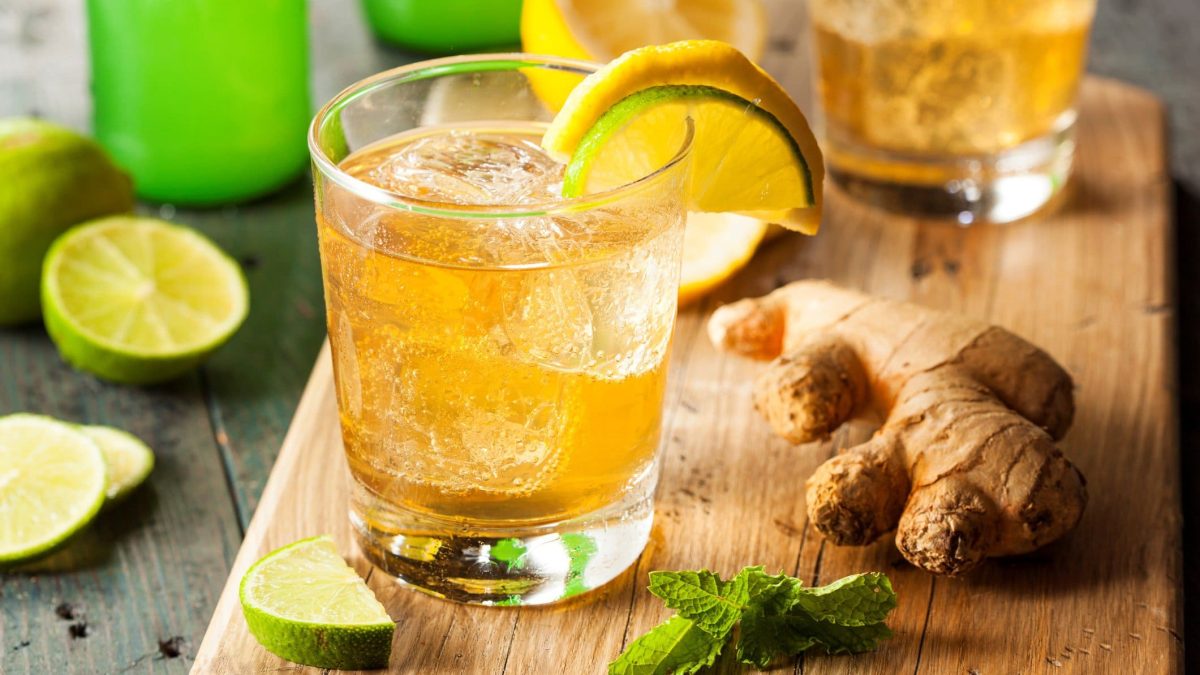 Zwei Ale Sangaree Biercocktails auf einem Holzbrett mit Zitronenmelisse, Ingwer und Limettenscheiben platziert auf einem Holztisch. Im Hintergrund weitere Limettenscheiben und Gläser angedeutet. Frontalansicht.
