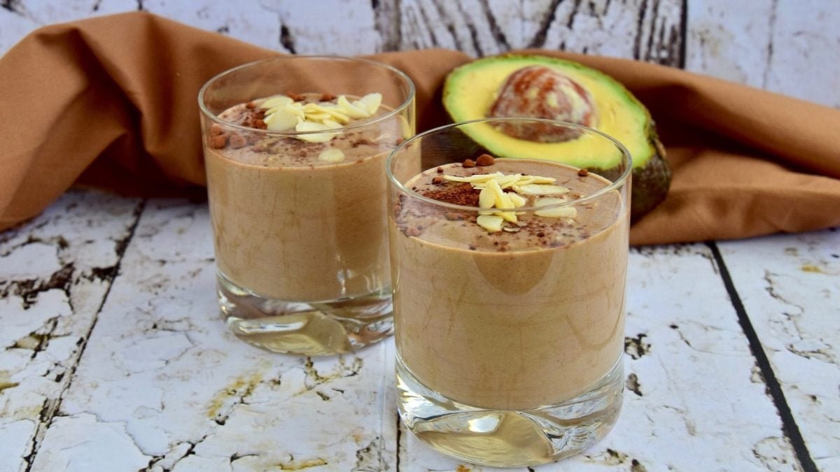 Zwei Gläser von unserem Avocado-Kakao-Smoothie stehen auf einem marmorierten Tisch. Im Hintergrund liegt eine halbe Avocado.