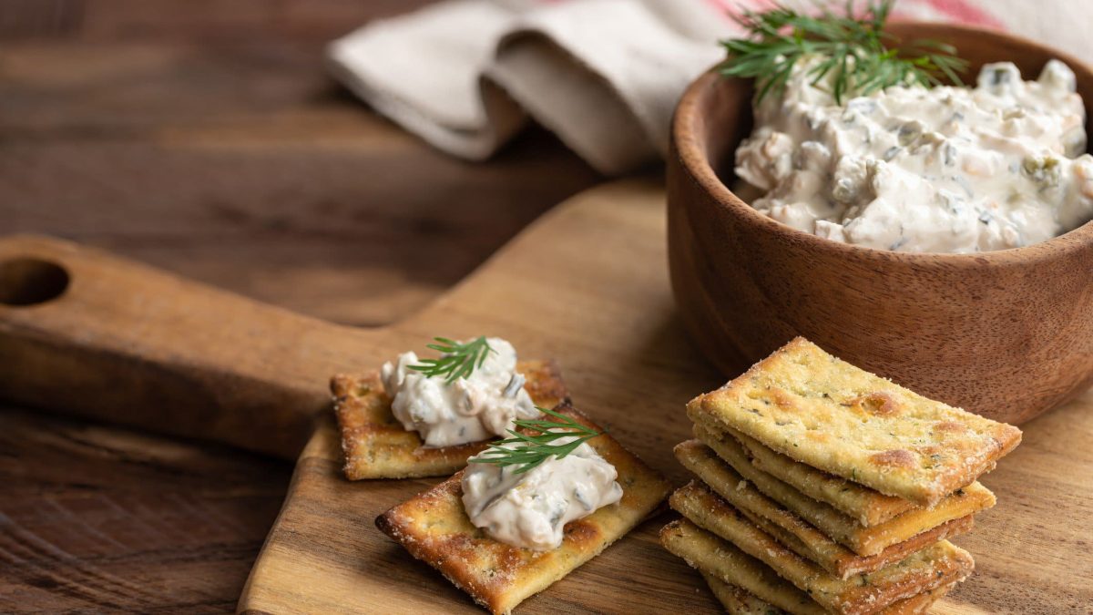 Bacon Onion Dip in einer Tonschale mit Crackern auf Holzbrett auf Holztisch. Weißes Tuch im Hintergrund. Frontalansicht.
