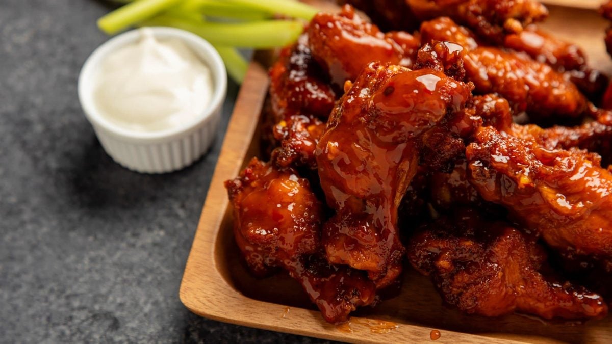 Blazin' Buffalo Wings auf Holzplatte angerichtet. Kleine Schale mit Creme Fraiche und Selleriestangen im Hintergrund. Platziert auf grauem Untergrund. Frontalansicht.