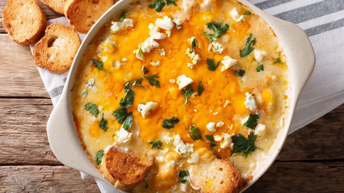 Buffalo Blue Cheese Dip in weißer Ofenform auf Tuch auf Holztisch. Auf dem Dip und daneben geröstete Brotscheiben. Aufnahme aus der Vogelperspektive.