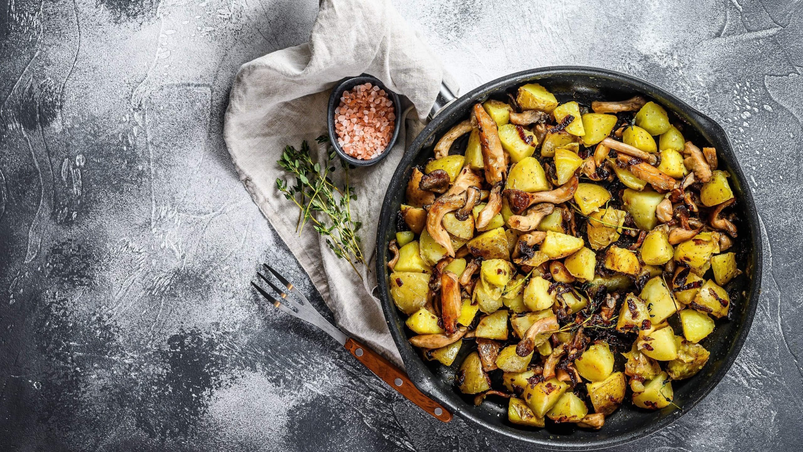 Rustikale gusseiserne Pfanne mit Bratkartoffeln mit Austernpilzen, garniert mit frischem Thymian auf einem bemehlten schwarzen Untergrund, von oben