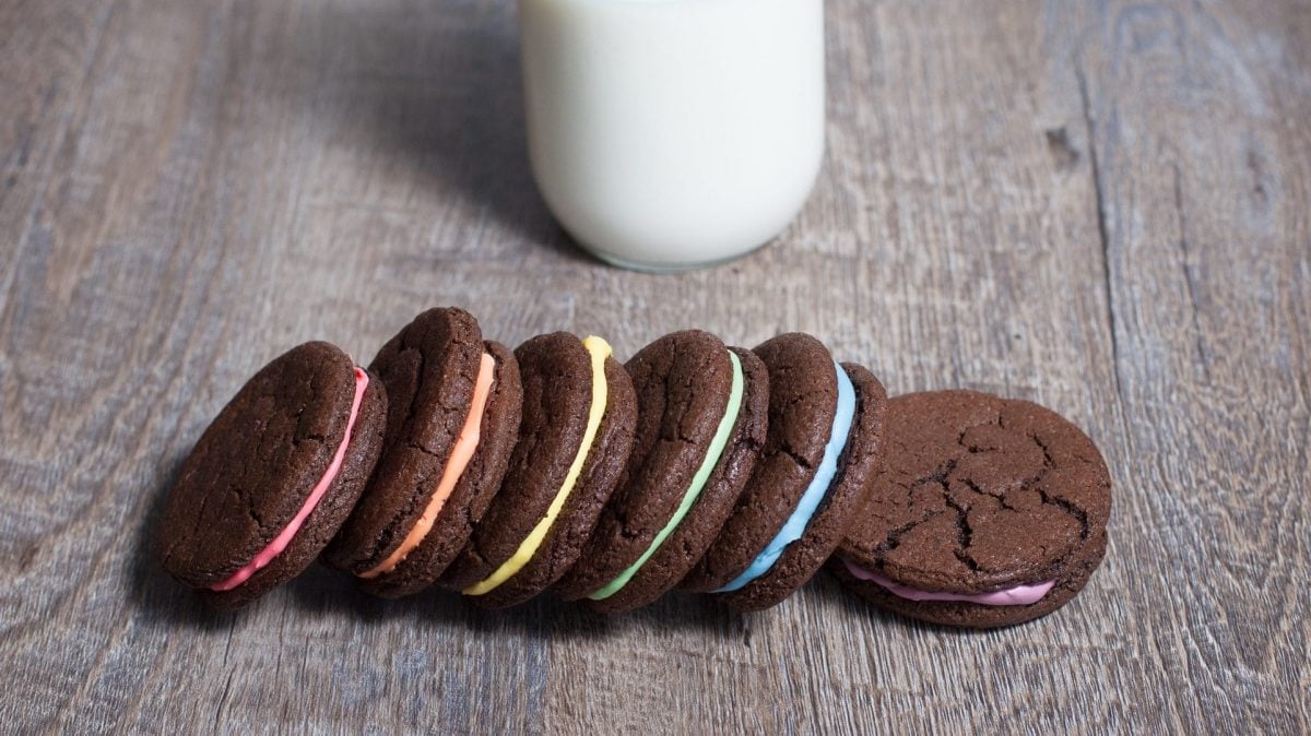Auf einem dunklen Holztisch liegen sechs bunte Oreos. Die Vanillecreme ist in den Farben des Regebogens. Im Hintergrund steht ein Glas Milch.
