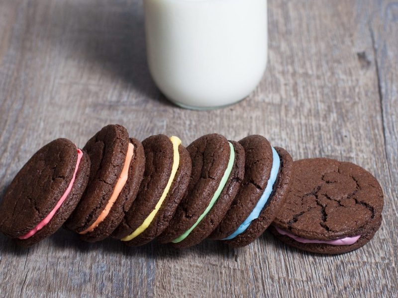 Auf einem dunklen Holztisch liegen sechs bunte Oreos. Die Vanillecreme ist in den Farben des Regebogens. Im Hintergrund steht ein Glas Milch.