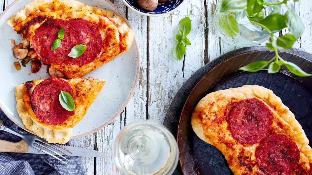 Calzone-Pfannenpizza auf einem Teller und in der Pfanne. Daneben Gläser mit verschiedenen Zutaten. Das Ganze auf einem hellen hölzernen Untergrund.