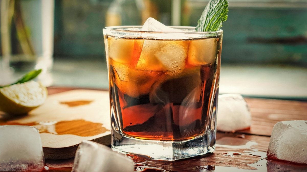 Cubeer Libre Bier-Cocktail mit Minze auf einem Holzbrett mit Eiswürfeln. Links Limettenviertel. Frontalaufnahme.