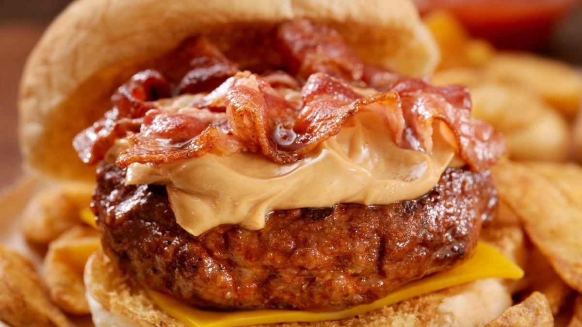 The Kings Finest Elvis Burger aufgeklappt mit Wedges auf Papier auf Holztisch. Im Hintergrund eine kleine Schlae aus Glas mit Ketchup. Frontalansicht