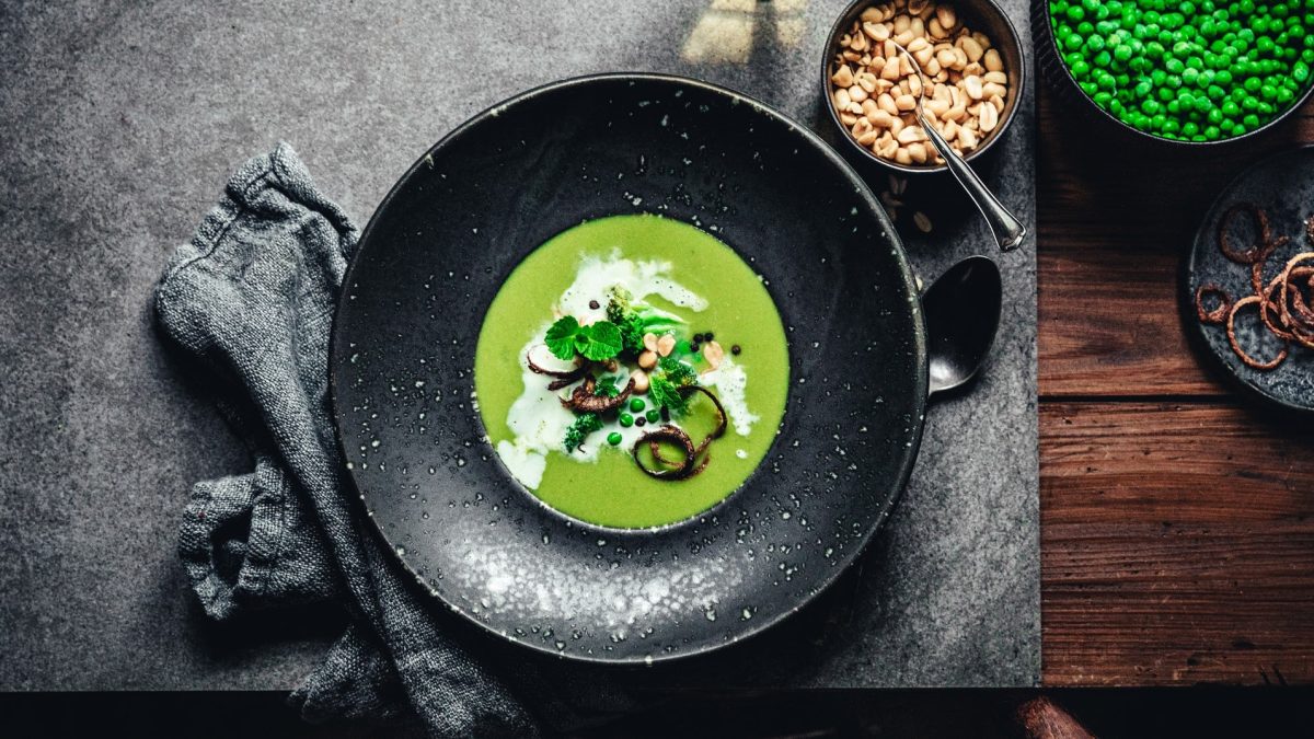 Ein schwarzer Teller Erbsen-Kokos-Suppe mit Minze, Erdnüssen und karamellisierten Zwiebelringen in der Draufsicht. Daneben stehen Schüsseln mit Zutaten.