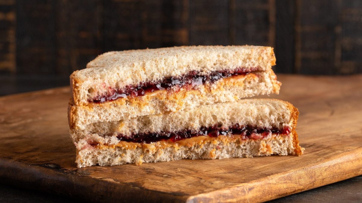 Ein Erdnussbutter-Marmeladen-Sandwich liegt halbiert und gestapelt auf einem Holzbrett.