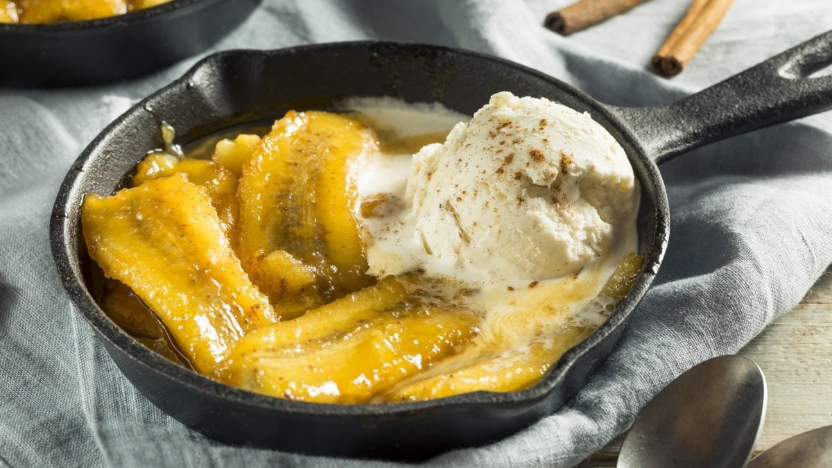 Flambierte Bananen mit Vanilleeis in einer gusseiserne Pfanne, mit Zimt bestreut auf einem grauen Tuch auf Holztisch