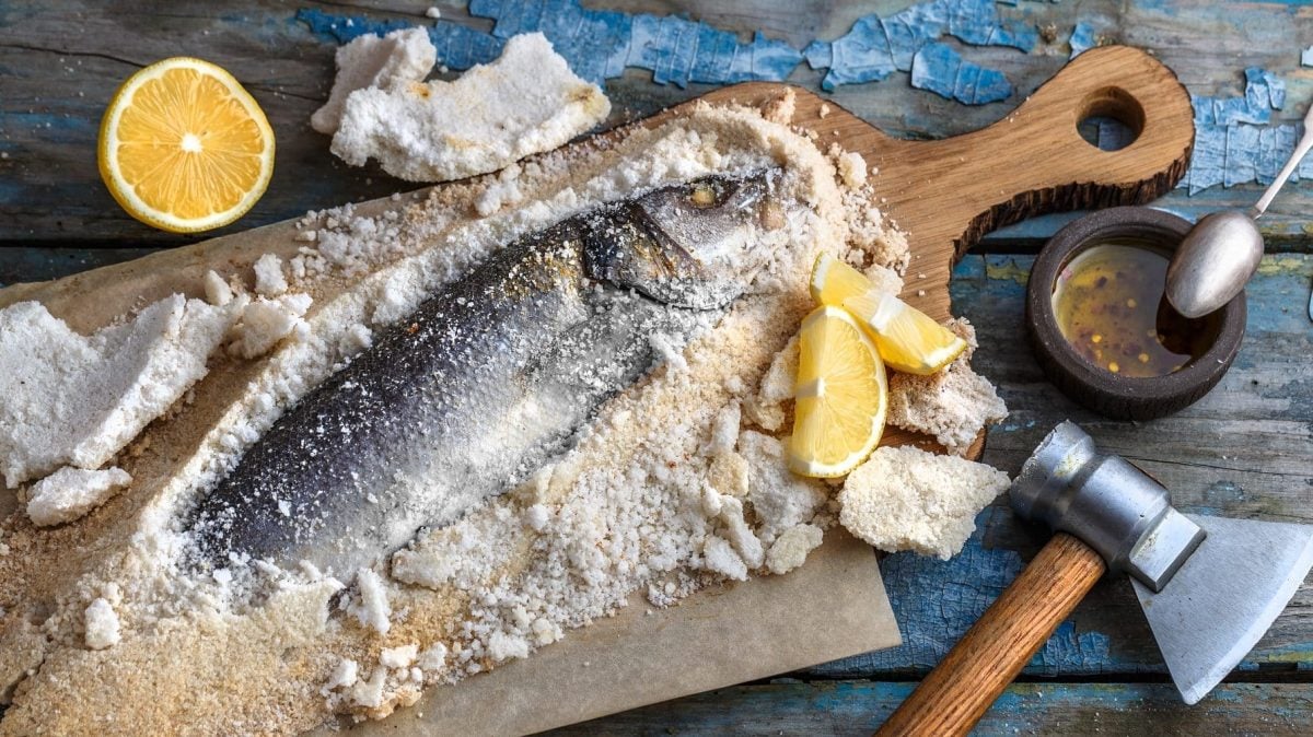 Aus der Vogelperspektive: Auf einem Holzbrett liegt ein Fisch in einer Salzkruste. Daben lieben ein Beil, Zitronenhälften, Salz und Pfeffer
