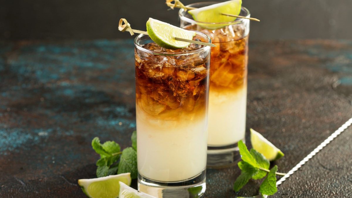 Zwei Frozen Espresso Dark and Stormy Cocktails in Gläsern mit Limettengarnitur auf Steintisch. Daneben Minzblätter, Limettenviertel und Metallspieße. Frontalaufnahme.