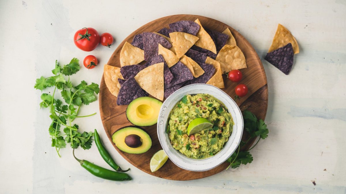 Guacamole auf rundem Holzbrett mit schwarzen und gelben Nachos, aufgeschnittener Avocado auf heller Steinplatte. Daneben Chile, Tomaten und Petersilie. Aufnahme aus der Vogelperspektive.