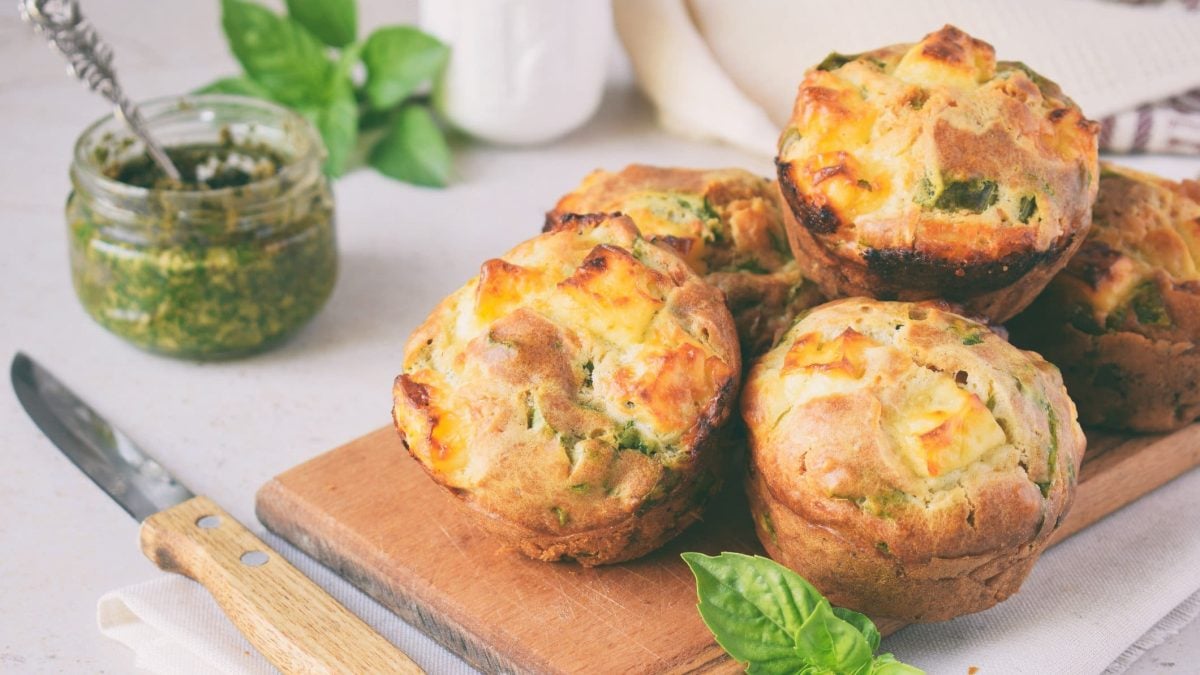 Glutenfreie Frühstücksmuffins auf einem Brett, daneben liegt ein Messer, außerdem ein Glas Pesto und einige Basilikumblätter.