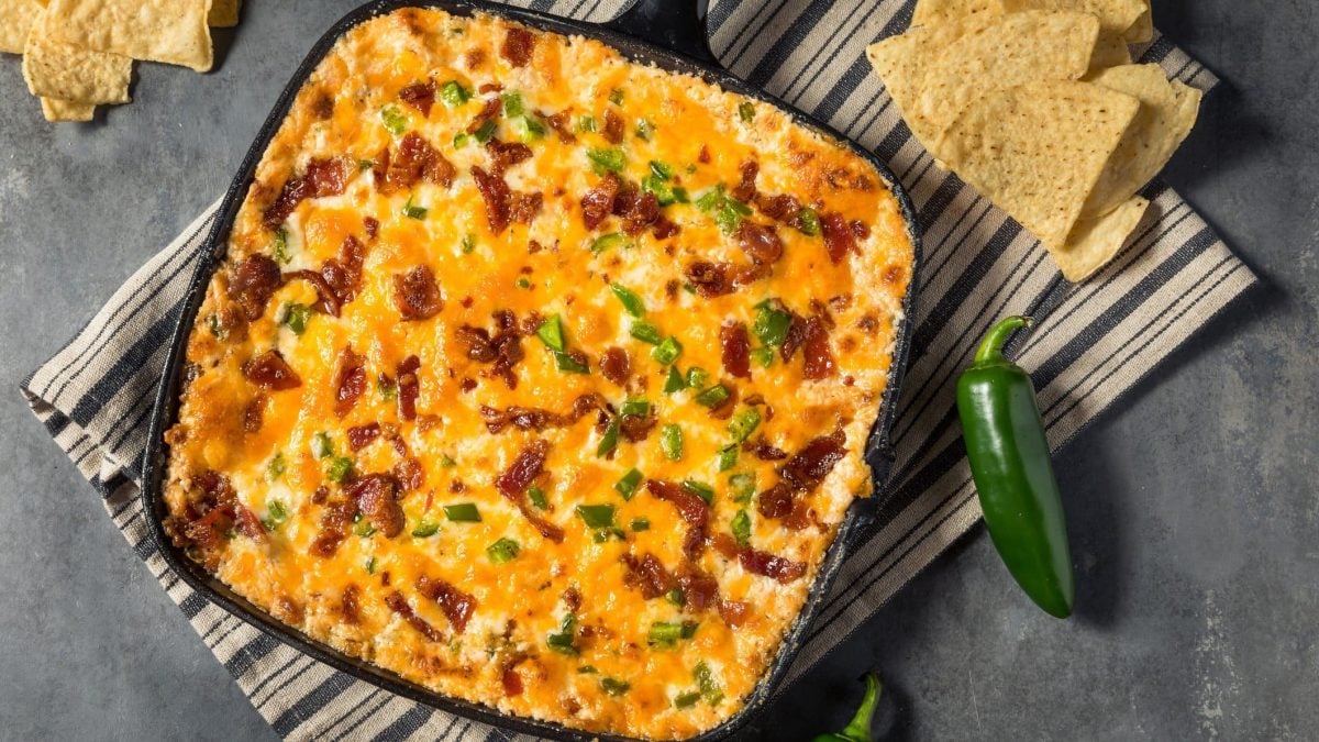 Jalapeño Popper Dip in Gusspfanne mit grüner Jalapeno und Nachos auf gestreifter Stoffserviette auf grauem Untergrund. Daneben Nachos und Jalapeños. Aufnahme aus der Vogelperspektive.