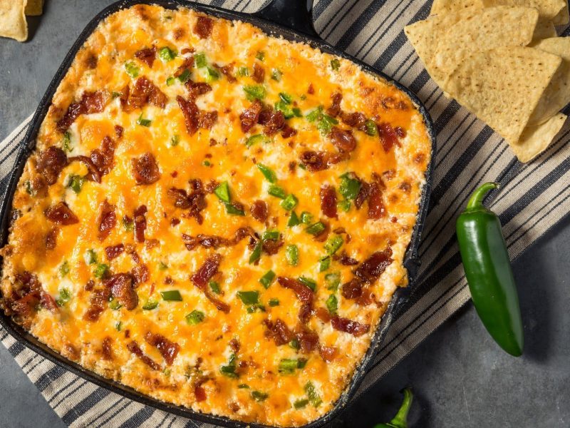 Jalapeño Popper Dip in Gusspfanne mit grüner Jalapeno und Nachos auf gestreifter Stoffserviette auf grauem Untergrund. Daneben Nachos und Jalapeños. Aufnahme aus der Vogelperspektive.