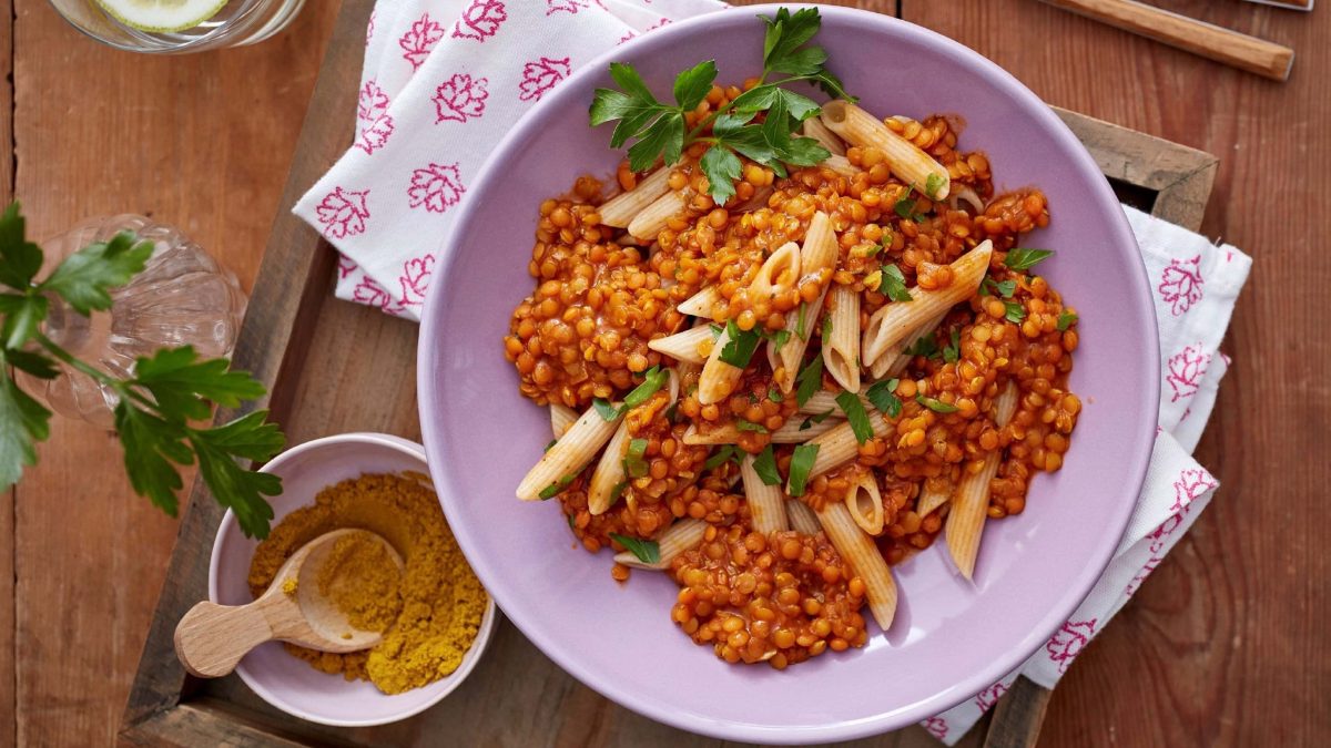 Linsen-Curry mit Pasta und frischer Petersilie in einer rosa Schale auf einem hölzernen Tisch