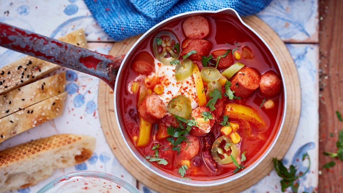 Ein mexikanisches Würstchen-Gulasch mit Paprika, Jalapeno, Tomaten, Würstchen und Crème Fraiche in einem Messingtopf auf einem Brettchen serviert