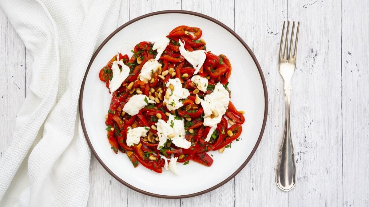 Aus der Vogelperspektive: Auf einem weißen Untergrund steht ein weißer Teller mit gerösteter Paprika mit Mozzarella und Pinienkernen. Daneben liegen ein weißes Tuch und eine Gabel.
