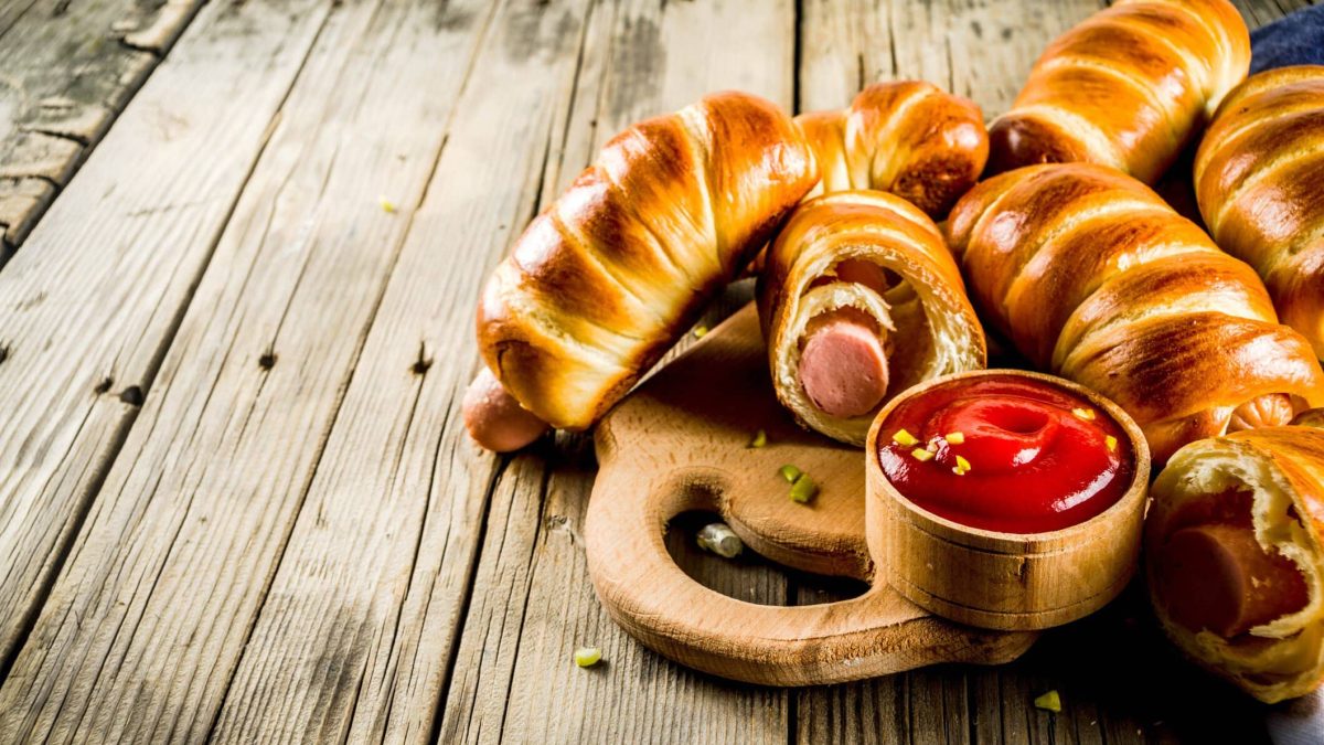 Pigs in Blankets zum Superbowl auf Holzbrett und Holztisch. Eines angeschnitten. Eine Schale mit Ketchup im Verdergrund und Würfel vo sauren Gurken als Deko verteilt. Frontalansicht.