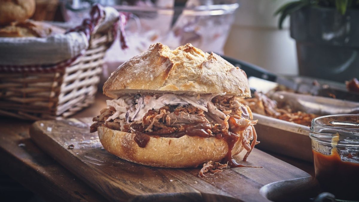 Ein Pulled-Pork-Sandwich mit Coleslaw auf einem Holzbrett. Im Hintergrund verschiedene Zutaten.