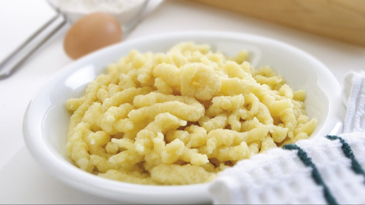 Ein weißer Teller mit selbstgemachten Spätzle steht auf einem Tisch. Daneben liegen ein Ei, eine Schale mit Mehl und ein Handtuch.