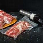 Sous-Vide Stab rechts und zwei vakuumierte Beutel mit rindfleisch links auf schwarzem Tisch