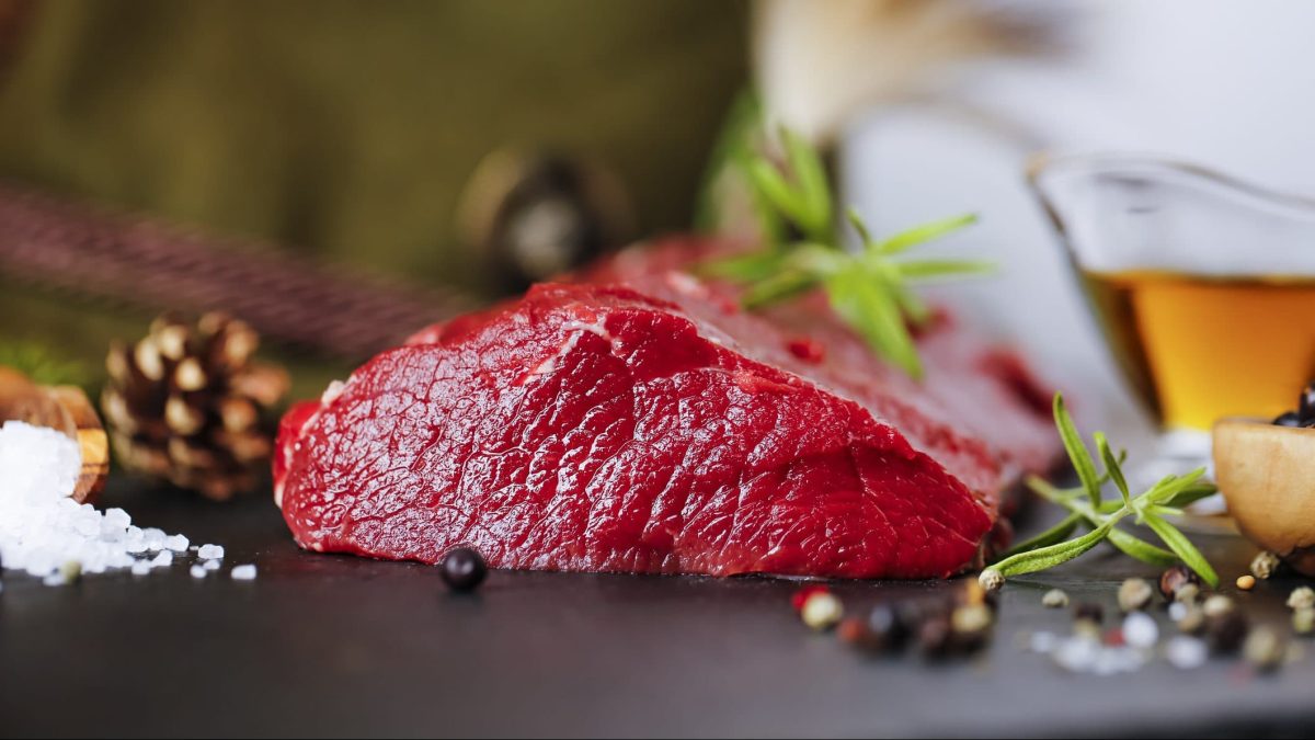 Auf einer dunklen Platte liegt Hirschfilet, aus dem Hirschsteaks geschnitten werden. Daneben liegt Salt, Pfeffer und Rosmarinzweige