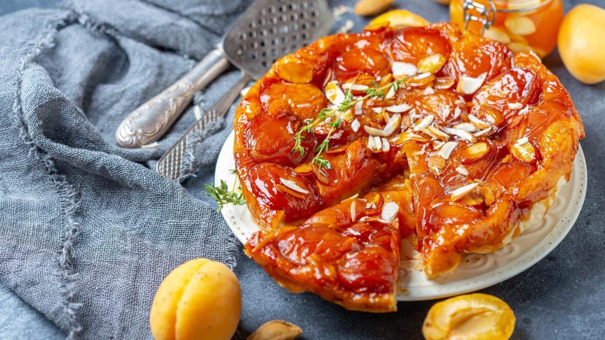 Tarte Tatin mit Aprikosen und Rosmarin angeschnitten auf einer Platte auf einem mit Stoff ausgelegtem Tisch, garniert mit frischem Rosmarin und Mandeln