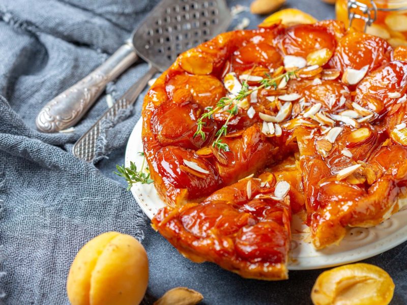 Tarte Tatin mit Aprikosen und Rosmarin angeschnitten auf einer Platte auf einem mit Stoff ausgelegtem Tisch, garniert mit frischem Rosmarin und Mandeln