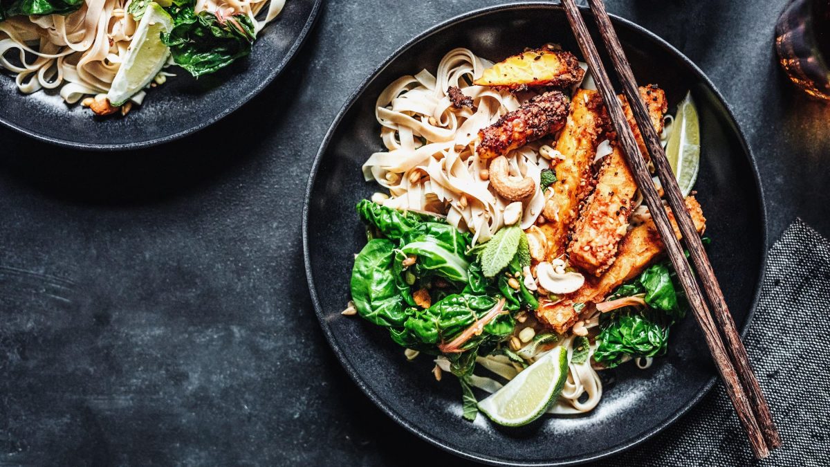 Tempeh-Spinat-Pfanne mit Reisbandnudeln, Kokosmilch, Limetten und gerösteten Nüssen in einer großen Schale auf Schieferuntergrund