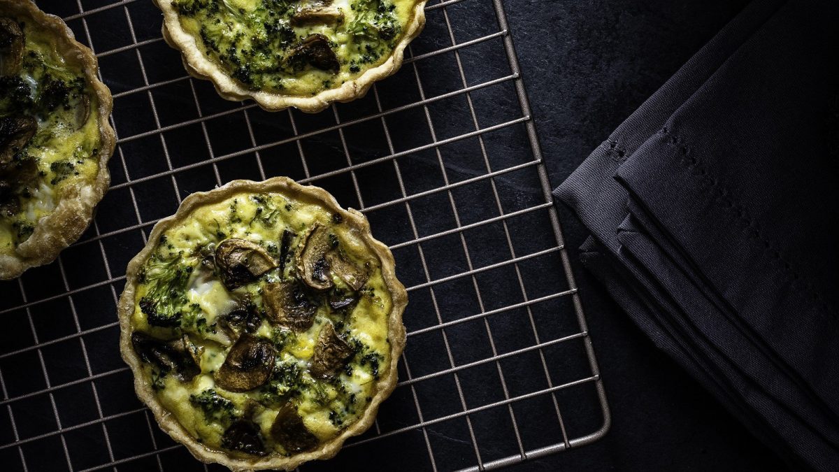 Saftige vegane Mini-Quiches mit cremiger Pilz-Brokkoli-Füllung auf einem Gitter vor schwarzem Hintergrund. Aufnahme aus der Vogelperspektive..