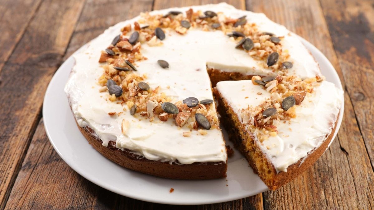 Veganer Karottenkuchen mit Frischkäse-Frosting und gerösteten Nüssen als Deko auf weißem Teller auf Holztisch plaziert
