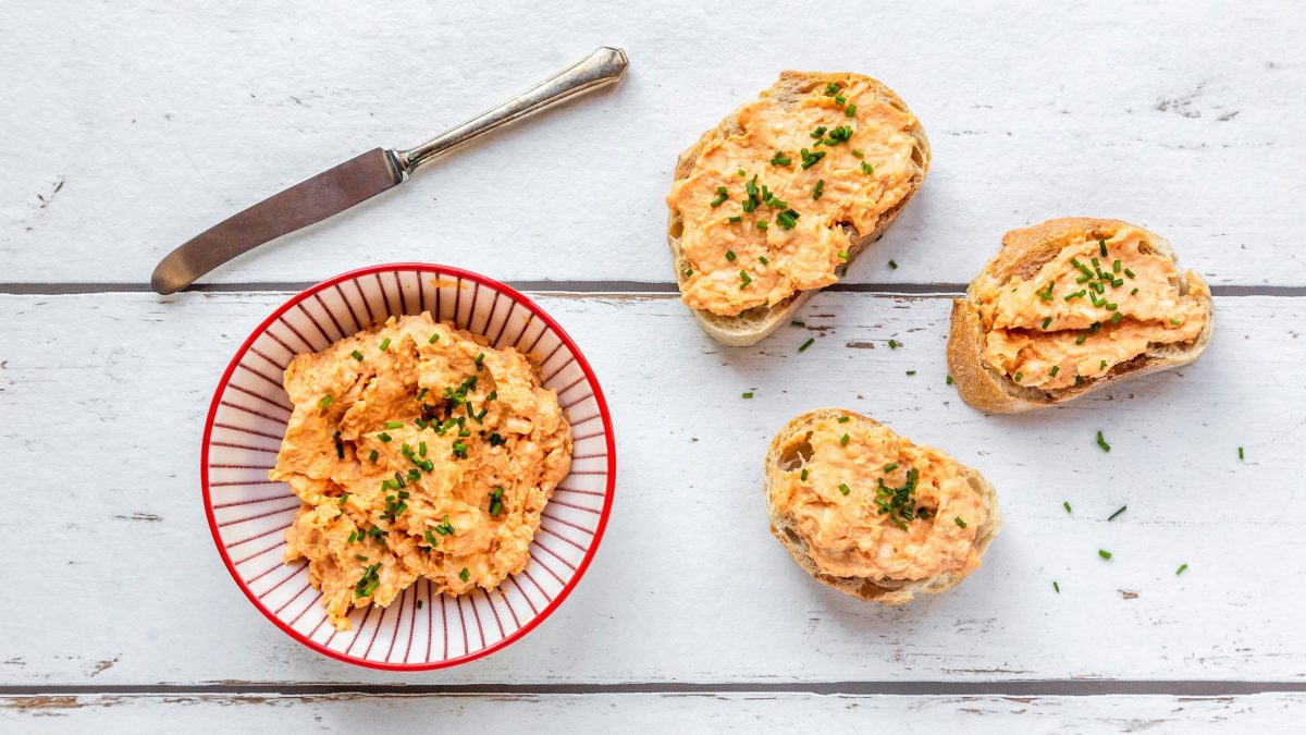 Eine Schüssel veganer Obazda mit einigen bestrichenen Baguettescheiben und einem Messer in der Draufsicht.