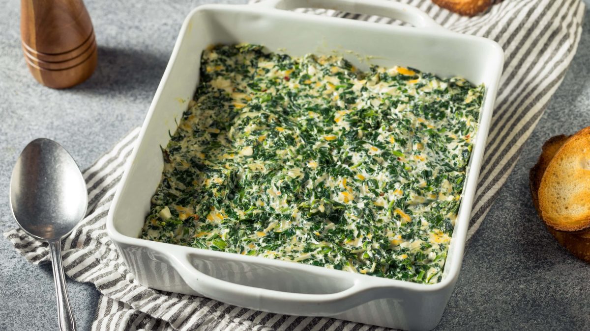 Eine Auflauffor, veganer gebackener Spinat-Artischocken-Dip, daneben ein Löffel und ein Stück getoastetes Baguette.