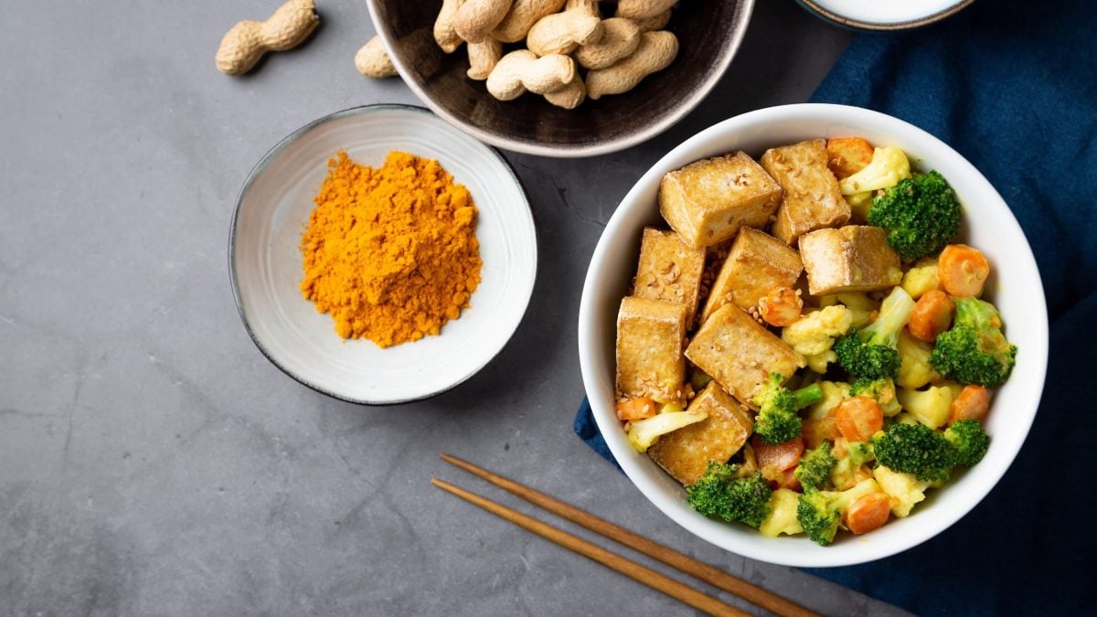 Wok-Gemüse in Erdnusssauce, bestehend aus Brokkoli, Blumenkohl, Karotten, serviert mit Tofuwürfeln in einer weißen Bowl auf dunklem Untergrund