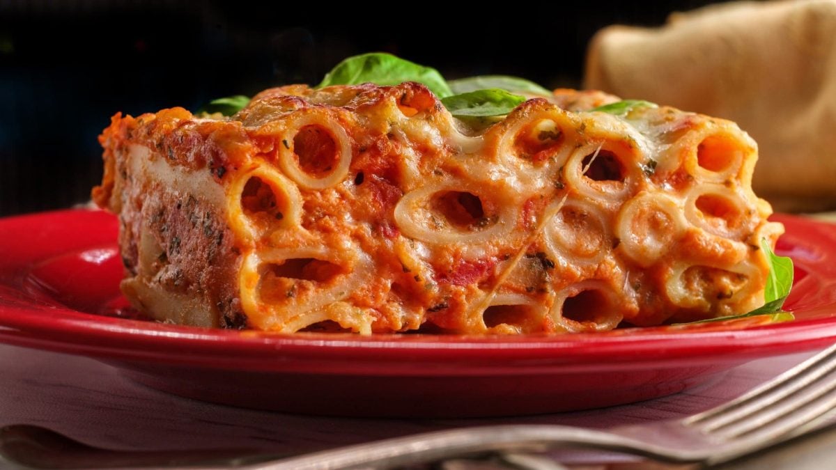 Ziti al Forno aus The Sopranos auf rotem Teller vor schwarzem Hintergrund. Dekoriert mit Basilikumblatt. Ein Gabel im Vordergrund. Frontalansicht.
