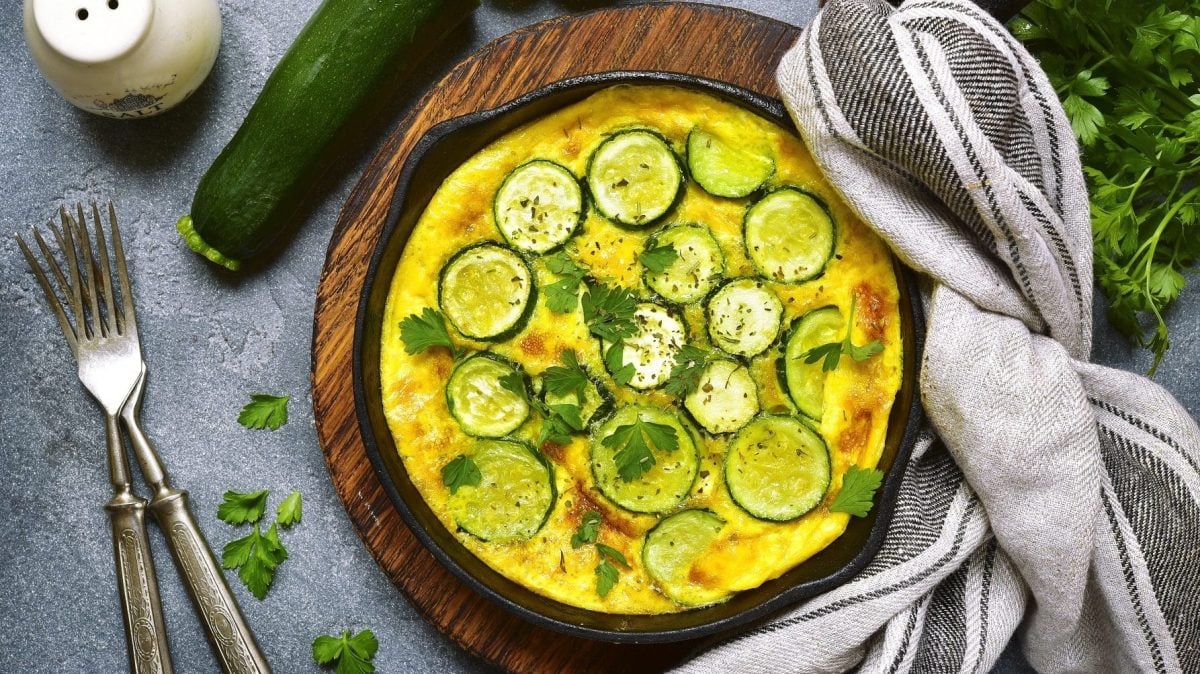 Aus der Vogelperspektive: Eine runde Zucchini-Frittata. Es handelt sich um eine Eierspeise. Neben der runden Form liegen ein handtuch, zwei Gabeln und frische Kräuter