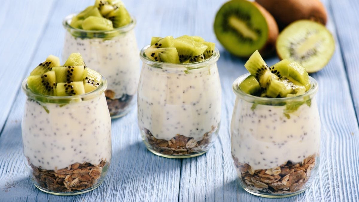 Drei Gläser Chia-Joghurt mit Müsli und Kiwi auf hellblauem Untergrund aus Holz.
