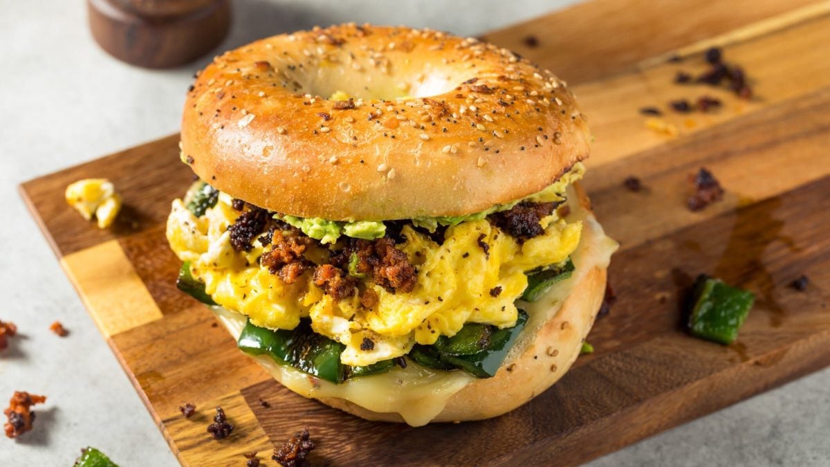 Bagel mit Rührei, grüner Paprika und Chorizo auf einem Holzbrett.