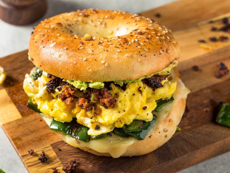 Bagel mit Rührei, grüner Paprika und Chorizo auf einem Holzbrett.