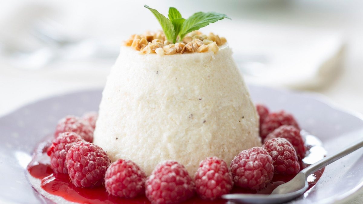 Auf einem Teller liegt ein Flummery Pudding. Er ist umrandet mit Himbeeren und mit gehackten Mandeln bestreut.