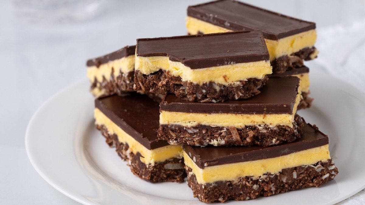 Auf einem Teller liegen mehrere Stücken selbstgemachter Nanaimo Bars. Sie bestehen aus einem Keksboden, einer Creme und einer Schokoschicht.