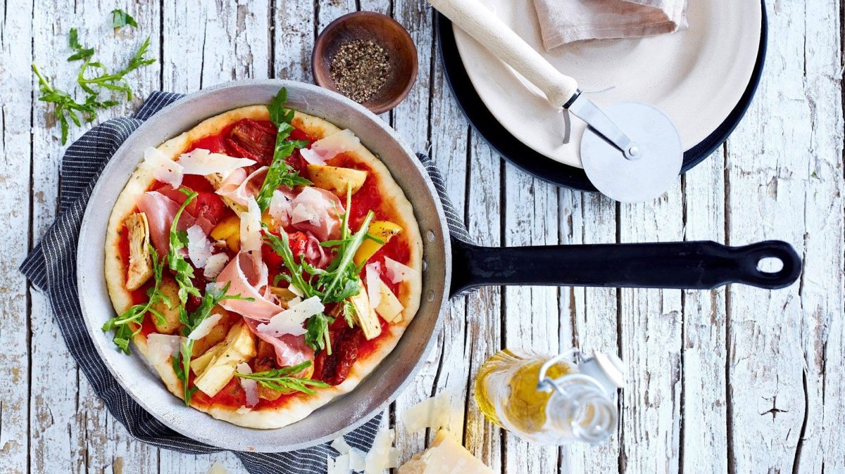 In einer Pfanne liegt eine fertige Pizza mit Antipasti. Sie ist verfeinert mit Schinken, Rucola und Parmesan.