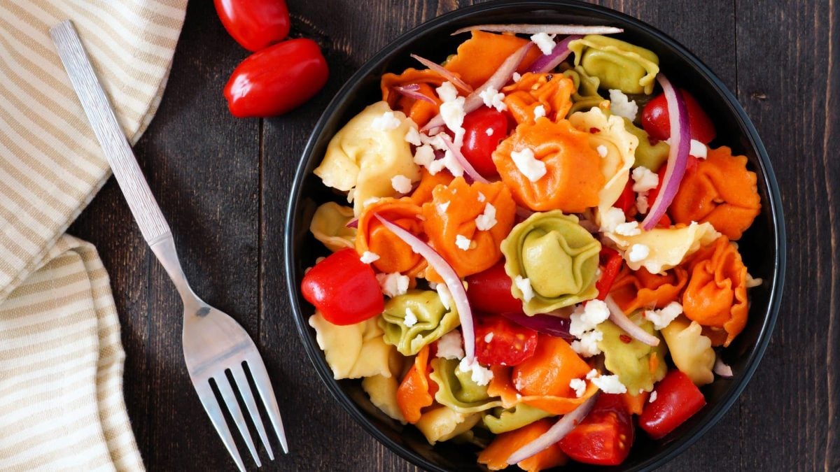 Unser bunter Tortellini-Salat steht angerichtet in einer Bowl auf einem braunen Holztisch.